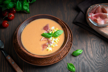 Salmorejo cold spanish soup made of pureed tomatoes and bread decorated with basil leaf and chopped boiled egg served in bowl with jamon or ham, spoon  on dark brown wooden table in summer. Horizontal