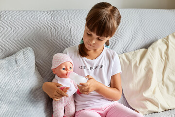 Indoor shot of little girl sitting on the sofa in the room at home playing with baby doll, like mom, dark haired preschooler female child holding toy.