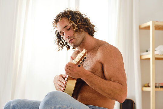 Shirtless man playing ukulele on bed at home
