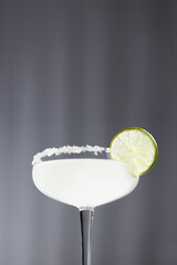Tropical fresh alcohol muddy white cocktail with salt rim and slice lime in hard light with striped shadows on white wall, copy space, vertical, closeup.