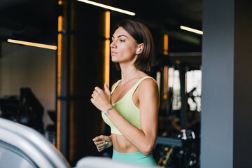 Fototapeta na wymiar Fit caucasian beautiful woman in fitting sport wear at gym running jogging at treadmill with wireless headphones in ears