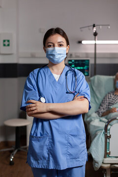 Portrait Of Medical Nurse With Chirurgical Mask In Hospital Room With Sick Unweel Senior Pantient With Trauma Looking At Camea With Arms Crossed, Wearing Scrubs.