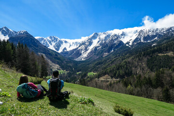 A couple with big hiking backpacks lying on a lush pasture and enjoying the panoramic view on Baeren Valley in Austrian Alps. The highest peaks are snow-capped. Lush green pasture. Clear and sunny day