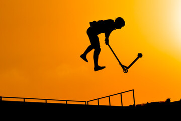 Unrecognizable teenage boy silhouette showing high jump tricks on scooter against orange sunsetwarm...