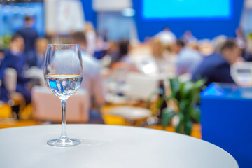 Close up view: glass of water, champagne or wine on white table against blurred business meeting, conference, seminar background. Corporate and communication concept