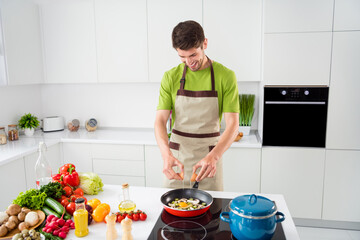 Portrait of attractive cheerful guy cooking tasty yummy healthy lunch dinner frying omelet at light white kitchen home indoors