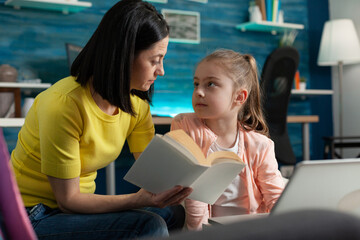 Mother assisting little child on reading school book while sitting at home. Smart girl with modern laptop on desk learning to read and studying for online classroom lesson lecture education