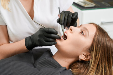 Female dentist in black sterile gloves examining patient teeth with dental explorer and mirror. Young woman lying in dental chair while having prophylactic examination. Concept of dentistry.