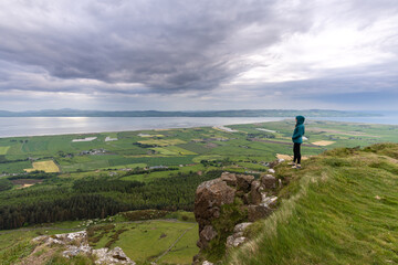 Caucasian female with a blue jacket standing in a green field with a beautiful view of the sea © Simon Izquierdo/Wirestock