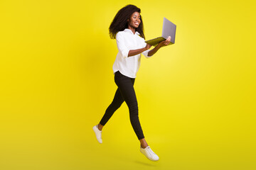 Full length photo of happy dark skin woman jump up walk hold laptop isolated on yellow color background