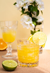 a crystal glass with a refreshing citrus drink or lemonade or alcoholic cocktail on beige flower background, sliced lime and orange are around goblets, minimalistic pastel still life