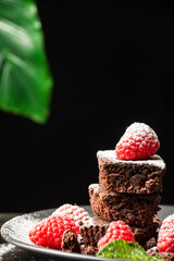 Close-up of plate with pieces of brownie, raspberries and mint leaves with sugar, selective focus, black background, vertical