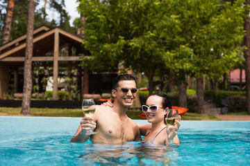 Cheerful couple in sunglasses holding glasses of wine in swimming pool outdoors