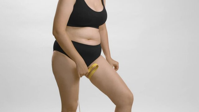 Full figured lady in black underwear wants to know her thighs volume using measure tape. White background still shot high quality studio video. Anonymous cowboy shot.