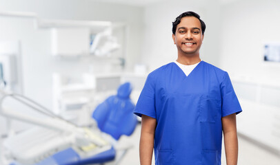 healthcare, stomatology and medicine concept - happy smiling indian doctor or male dentist in blue uniform over dental clinic office background