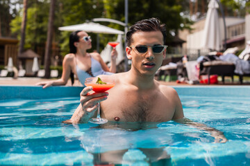 Man with wet hair holding cocktail in swimming pool near blurred girlfriend