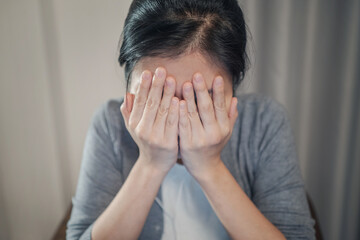 A close-up shot of a stressed and sad young adult Asian woman, in a gray cardigan and white t-shirt inside. She covering face with her hands.