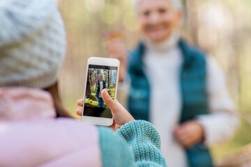 picking season, leisure and people concept - granddaughter with smartphone photographing happy smiling grandmother with mushrooms in basket in forest