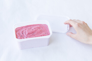 Berry ice cream pink color cream texture in a rectangular white container with woman hand is opening a lid on a white background. Top view