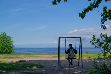 An adult guy, a man in a cap and with a backpack sits on a swing and sways among the trees against the background of the blue sea and sky. Back view