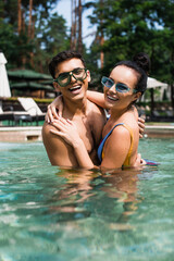 Obraz premium Surface level of positive couple hugging and looking at camera in blurred swimming pool