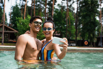 Smiling woman embracing boyfriend while taking selfie in swimming pool on resort
