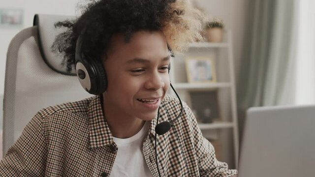 Chest-up of energetic African teenager wearing headset, sitting at desk in his room, talking on microphone and failing in online game