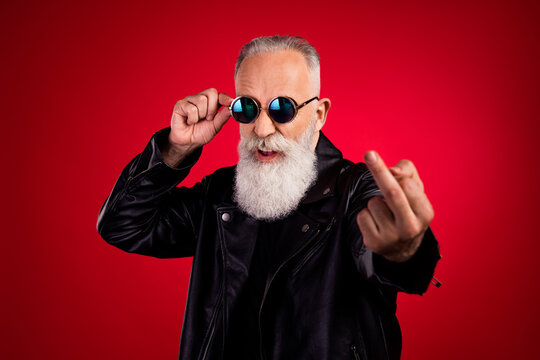 Portrait of attractive fashionable elderly grey-haired man showing middle finger touching specs isolated over bright red color background