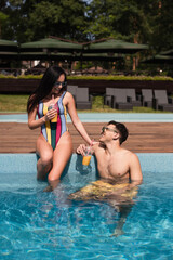 Cheerful woman with orange juice sitting on poolside near boyfriend in water