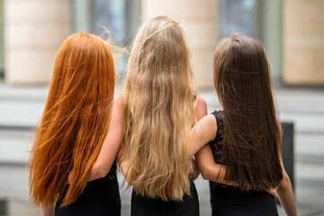 In summer, three teenage girls with loose long hair, a blonde, a redhead and a brunette, stand on...