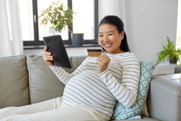 Obraz premium pregnancy, online shopping and people concept - happy smiling pregnant asian woman with tablet pc computer and credit card sitting on sofa at home