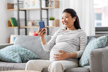 pregnancy, rest, people and expectation concept - happy smiling pregnant asian woman with smartphone and earphones sitting on sofa at home and listening to music