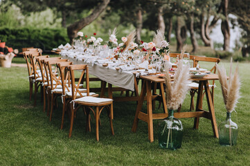 A large, long, decorated, wooden table and chairs, covered with a white tablecloth with dishes, flowers, candles, stands on the green grass in the park, in the forest in nature. Wedding banquet.