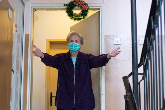 An Old Grandma Standing In Front Of Apartment Waiting For Her Grandkids To Visit Her. Hug Me Photo Of Grandma Spread Open Arms Wear Mask. Blue Eyes And Grey Hair