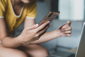 Female hand holding smart phone and credit card sitting on sofa at home to pay for items through an online payment system. Online shopping is the best option to reduce the spread of the coronavirus.