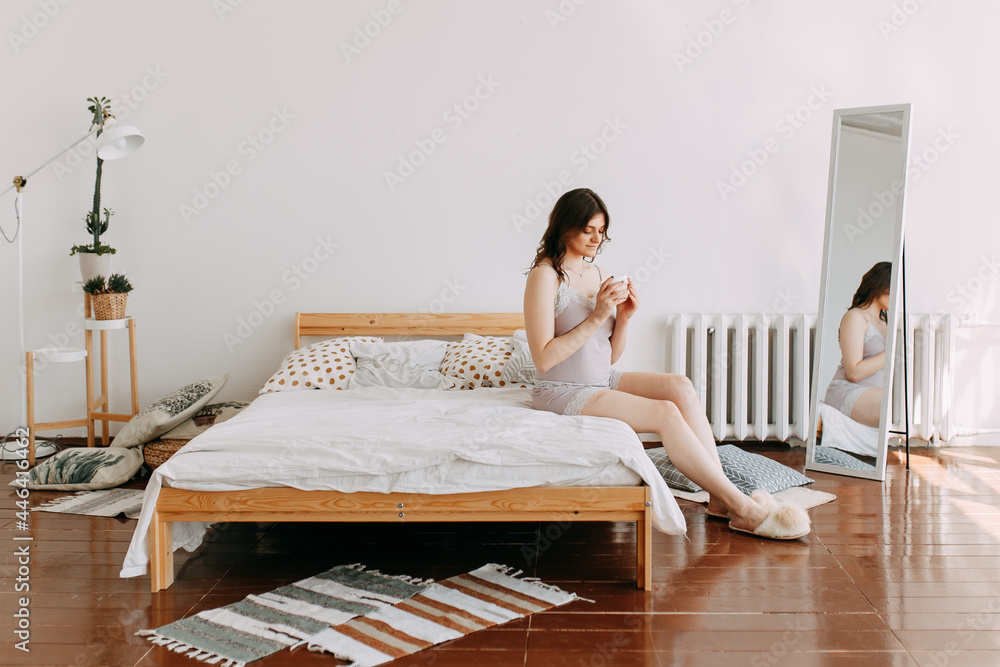 Wall mural a young smiling sleepy teenage girl is resting at home drinking coffee holding a mug sitting in bed 