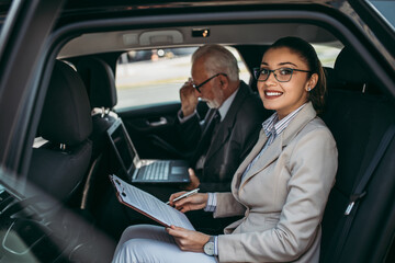 Good looking senior business man and his young woman colleague or coworker sitting on backseat in...