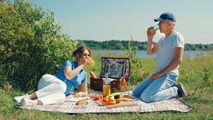 A slender elderly couple is relaxing on the grass in a park by the river and enjoying fruit and...