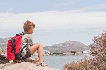 a boy with a backpack looks into the distance at a port at sea