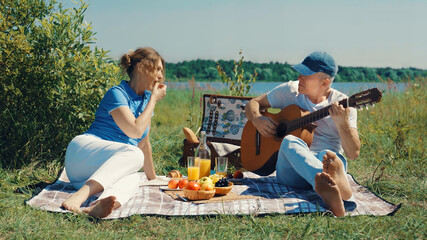 A happy elderly slender couple, a man and a woman, relax with a guitar on a picnic on the river...