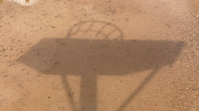 Der Schatten eines Basketballkorbs, in einer Regenpf&uuml;tze, auf sandigem Boden.