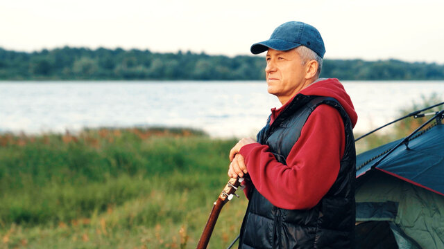 An elderly male tourist with an acoustic instrument enjoys nature and the sunset sunlight. Lifestyle, the concept of rest in old age. An elderly slender man with a guitar by the river at sunset.