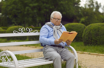 Elderly senior man wearing eyeglasses enjoy reading book sitting on wooden bench in city public park outside. Happy retirement lifestyle, hobby and pleasure. Concept of serene old age