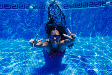 happy caucasian woman wearing sunglasses diving in swimming pool. underwater view. making V sign. Summer time and vacation concept