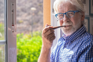 Smiling senior man at the window looking at camera while smoking a pipe