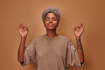 Mindful peaceful African American woman meditates indoor keeps hands in mudra gesture has eyes...