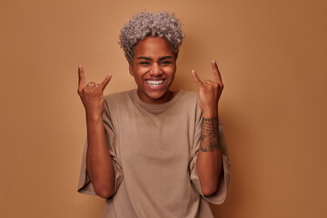 Positive joyful dark skinned woman makes heavy metal sign has fun on music rocking festival exclaims loudly closes eyes gestures actively, dressed in casual wear isolated over brown wall. Rock n roll.