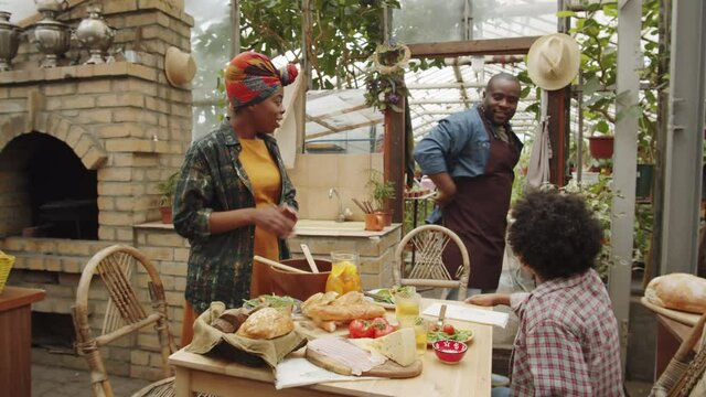 Afro-American Dad And Son Walking To Table While Mother Putting Food On Plates And Chatting With Them At Family Dinner In Greenhouse