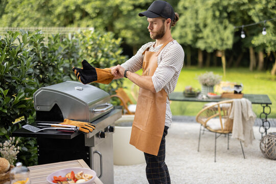 Handsome Man In Leather Apron And Gloves Preparing For Cooking On The Modern Gas Grill At Backyard Of Country House