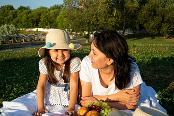 Mom and daughter in the park are lie on the grass on a white blanket, dressed in white clothes. They eat fruits and berries. Family on a picnic.